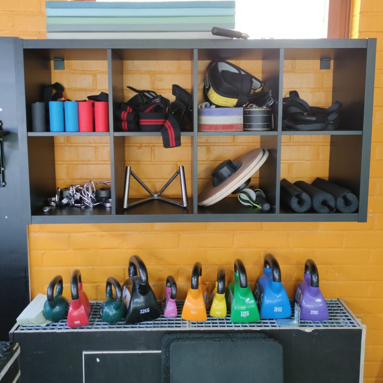 Colourful kettlebells and a shelf with lifting belts, wrist wraps and a balance board