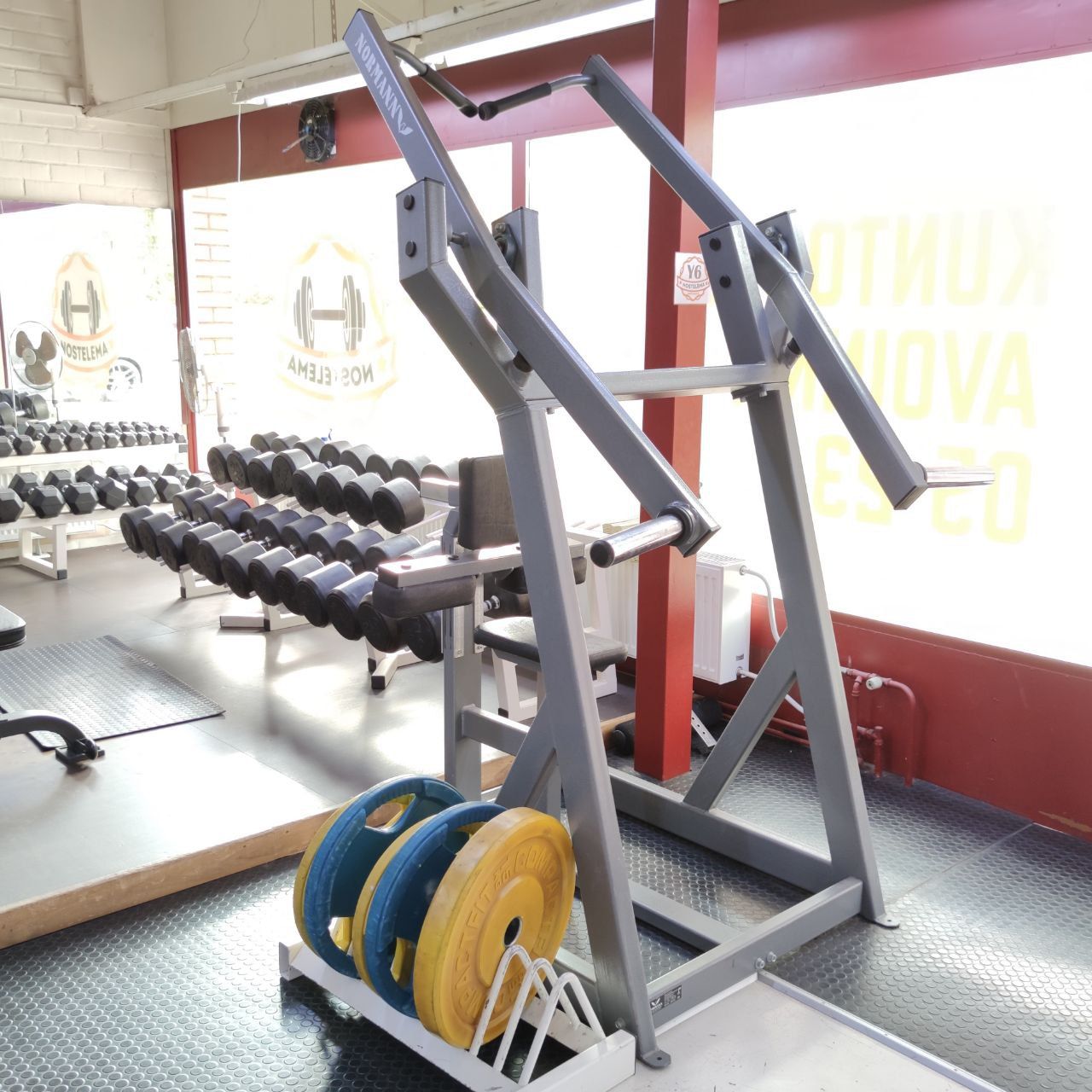 Plate-loaded lat pulldown with lever arms and a seat, with dumbbells in the background
