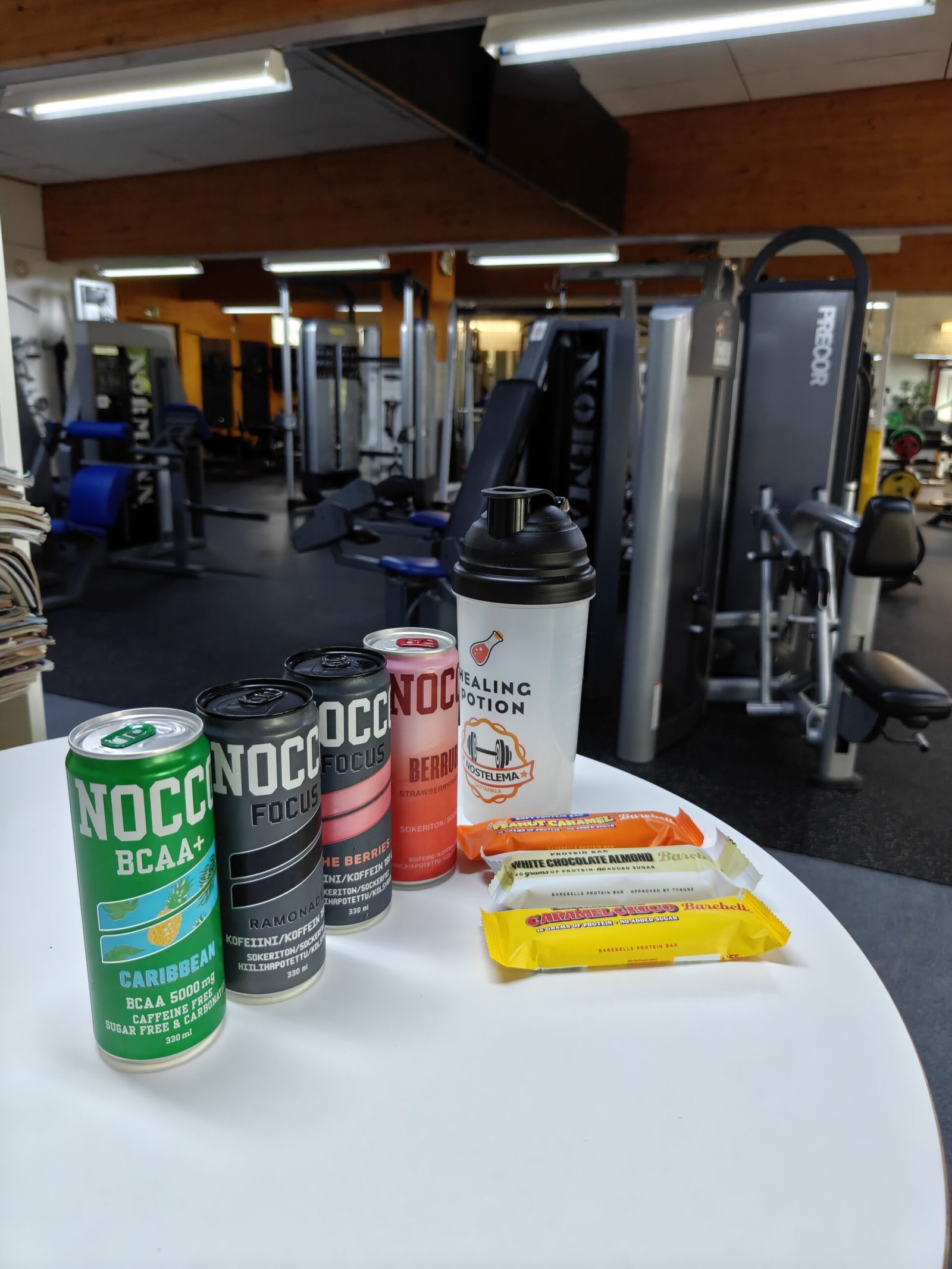 Nocco cans, Barebells protein bars and a Nostelema Healing Potion shaker on a table at the gym
